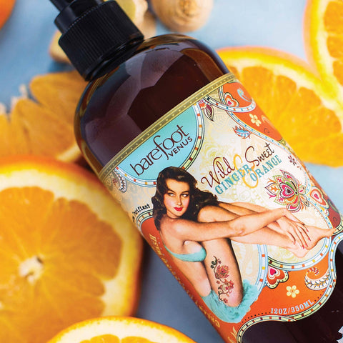 Bottle of 'Wild & Sweet Ginger Orange' body wash with a colorful label featuring a vintage-style model, surrounded by sliced oranges.