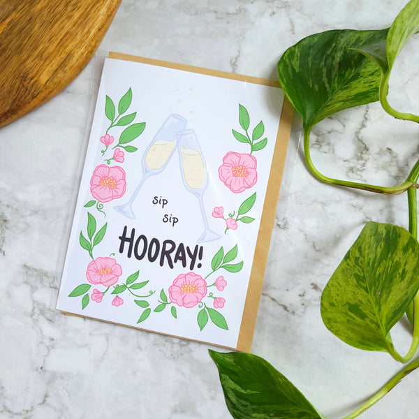 A 5x7 inch greeting card with a floral design and the text 'sip sip hooray!' displayed. The card is placed on a marble surface with a green leafy plant beside it.
