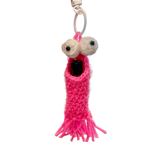 Pink knitted keychain with googly eyes held in a hand against a blurred store background