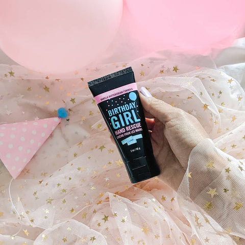 Hand holding 'Birthday Girl Hand Rescue' cream against a festive background with pink balloons and streamers.