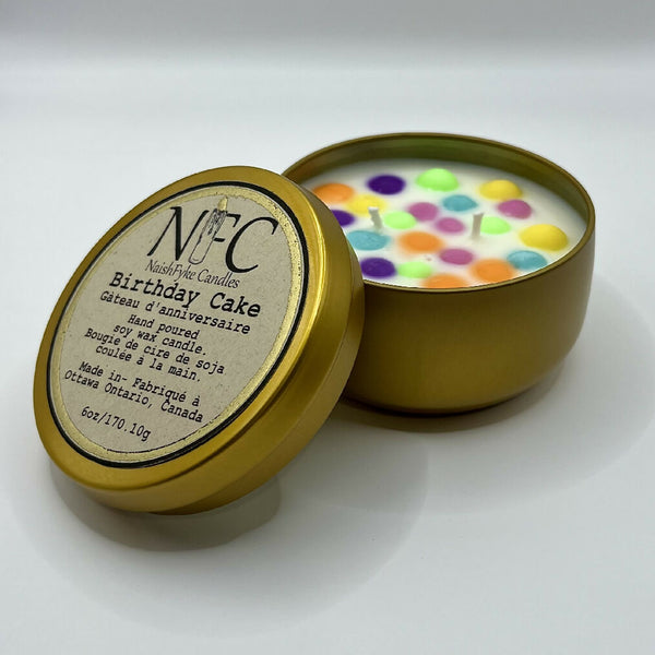 A gold colored candle tin with a lid, labeled 'Birthday Cake', containing a white soy wax candle with multicolored dots on top.