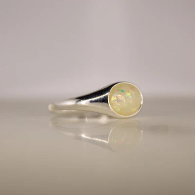 Silver ring with a round opal stone on a reflective surface with a light gray background