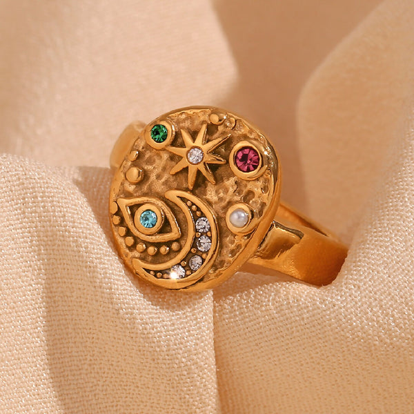Gold ring with star moon and eye designs and gemstones on a beige fabric background